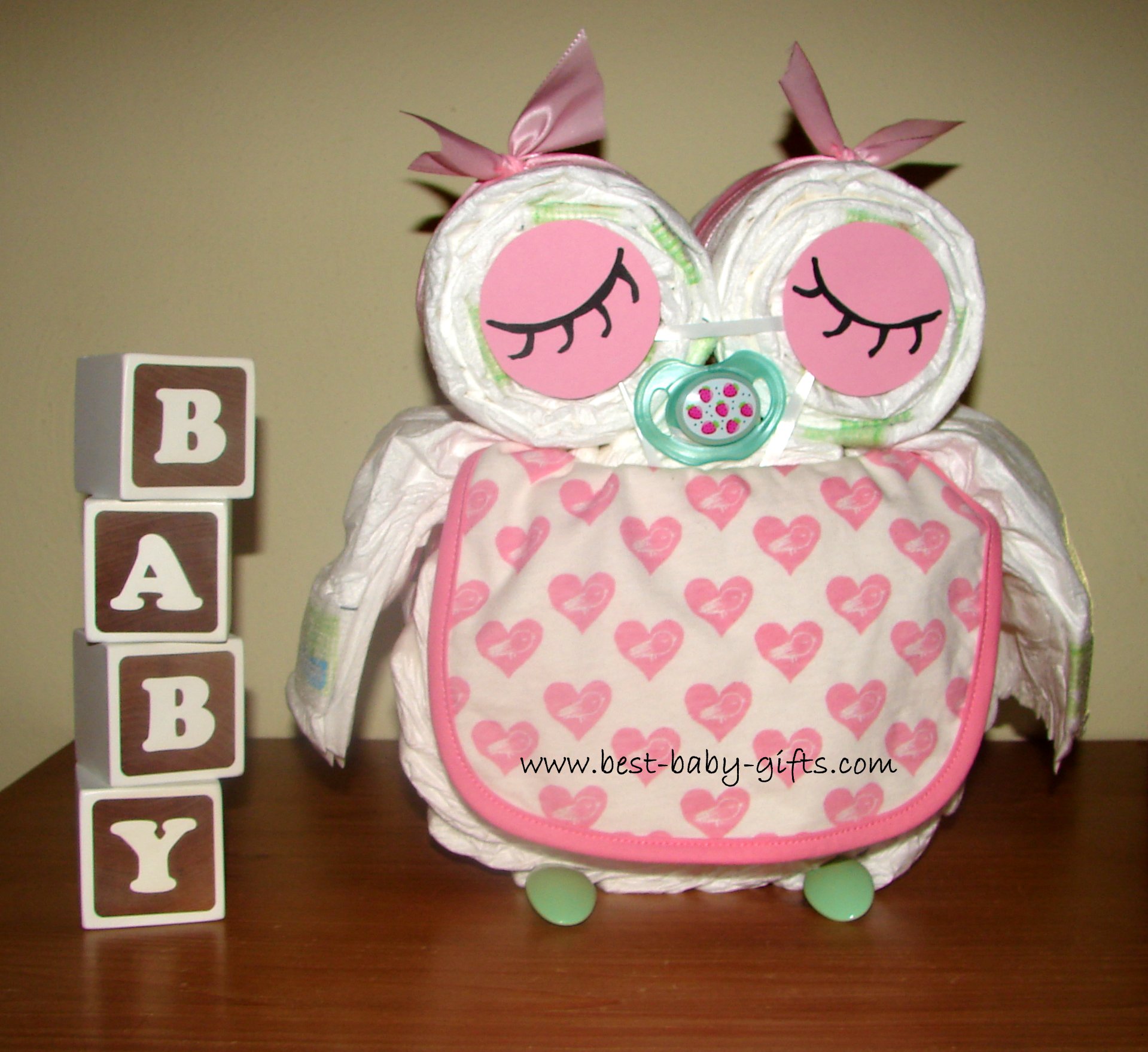 rosa Windeleule mit grünen Akzenten, mit Herzchenlatz, links davon 4 aufgetürmte weiße Spielklötze aus Holz "Baby"