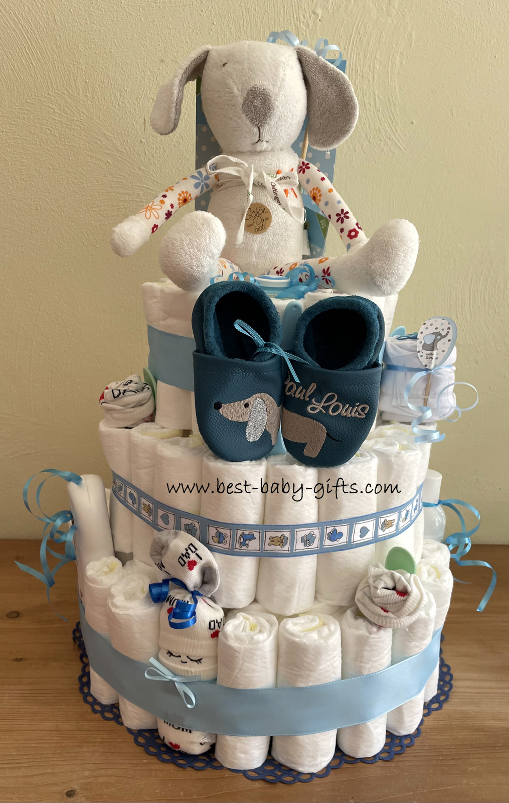 a large diaper cake with a cuddly dog on top, a pair of blue baby shoes and a diaper baby at the base layer, blue details