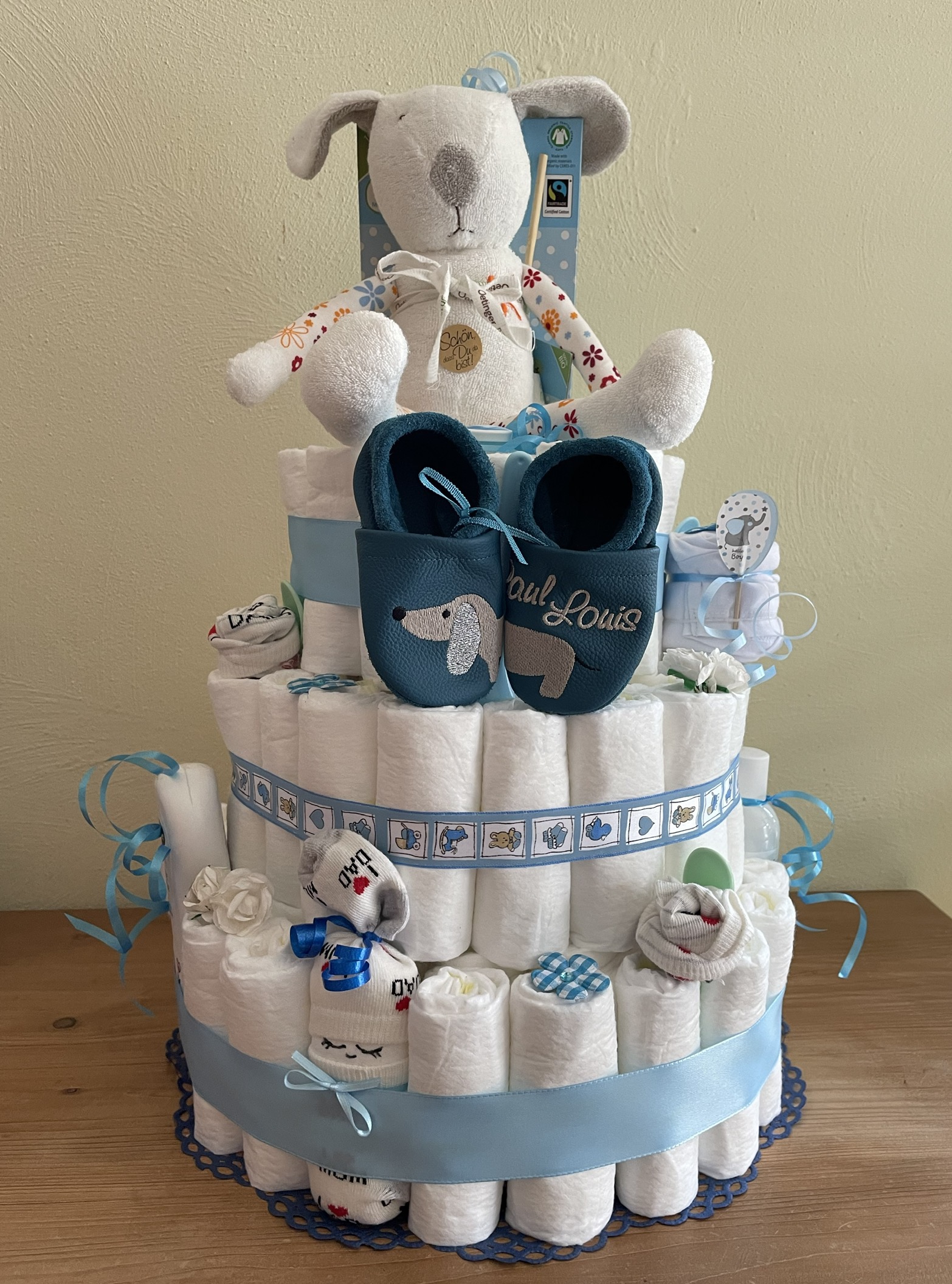 a 3-tier diaper cake for a baby boy in blue featuring a pair of leather booties with a dachshund and baby's name on and a large, cute dog toy for baby on top of the cake