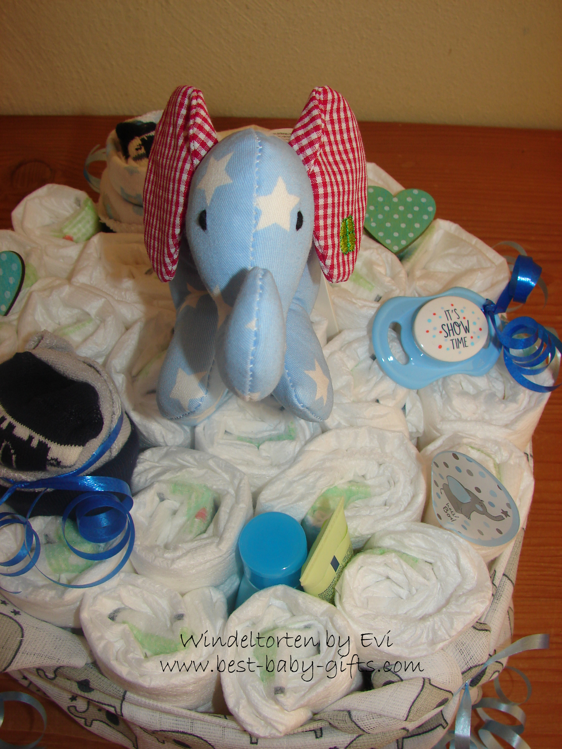 a one tier diaper cake shown from above, a blue cuddly elephant is in the middle and decorations are a pacifier, socks and you can also see some bottle caps
