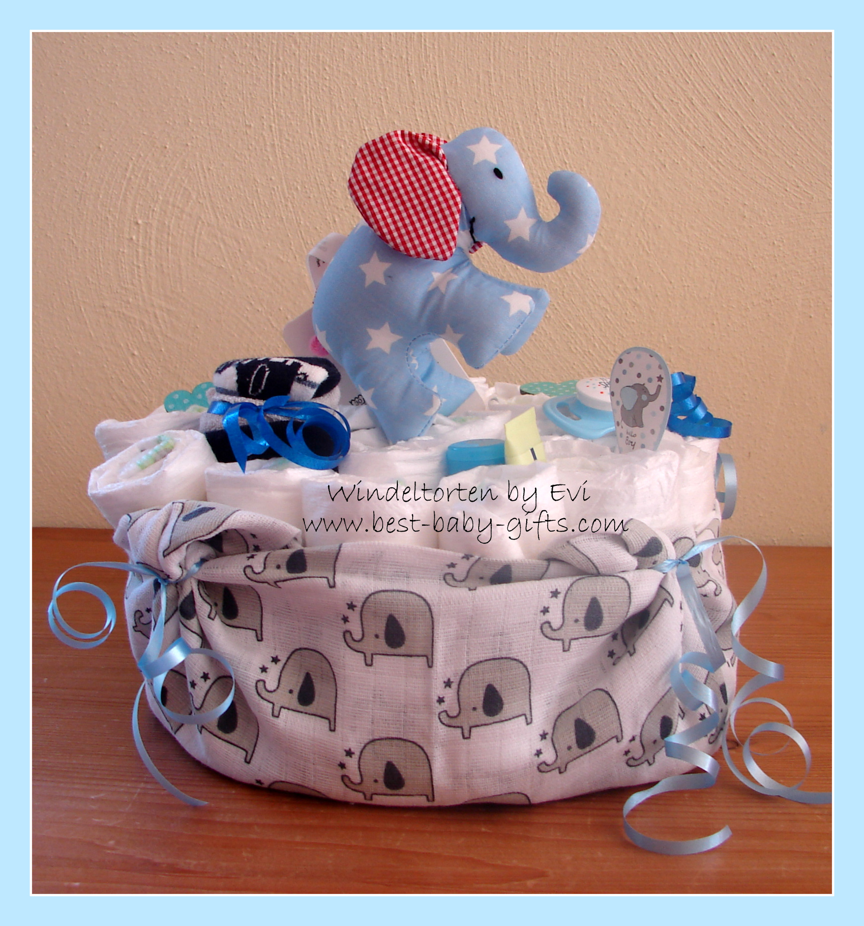 a round one tier diaper cake wrapped in a cloth with elephants all over design, on top of the diaper cake is a blue cuddly elephant with red ears