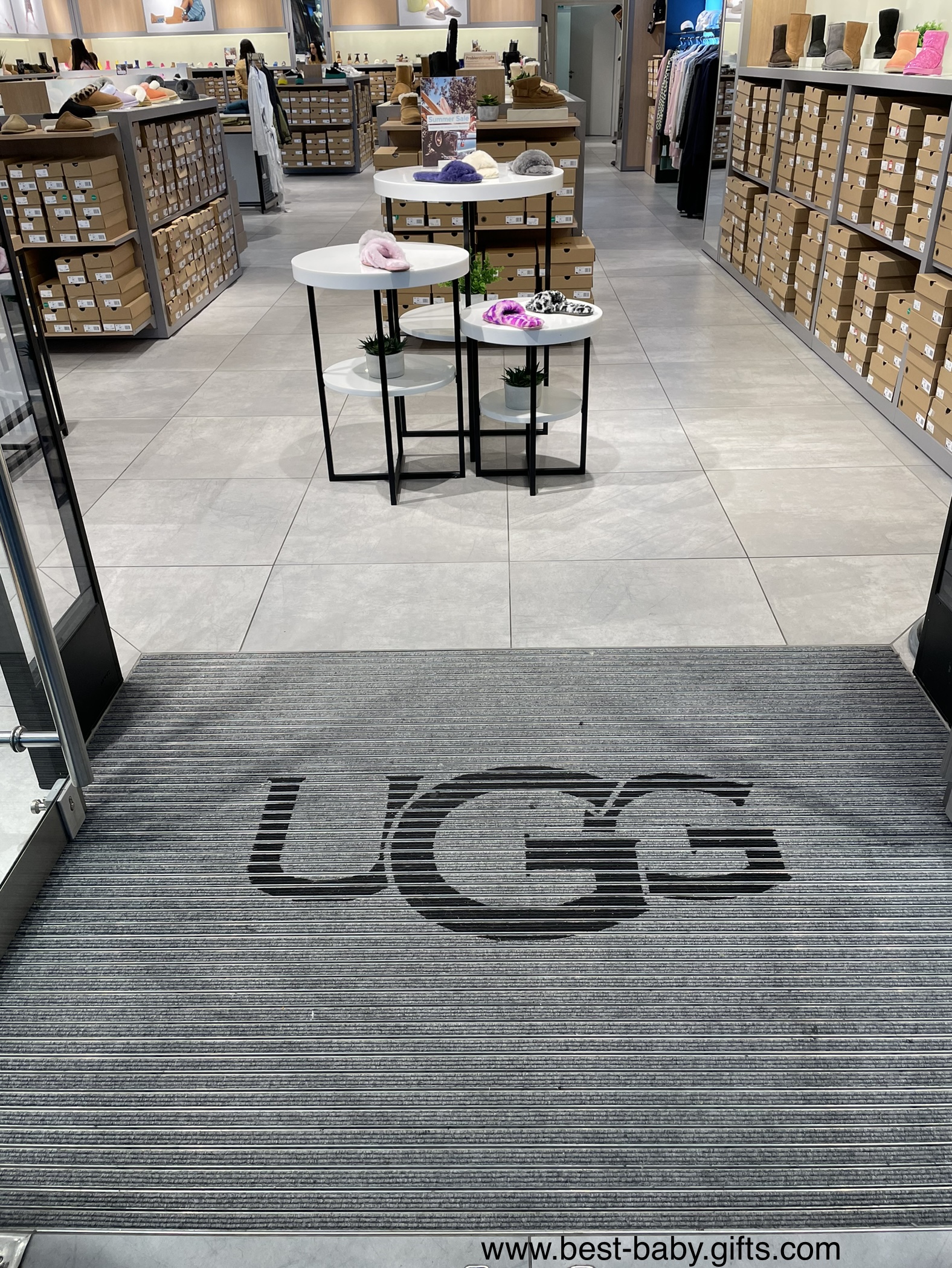 a modern UGG store entrance, doors wide open, it says UGG on the floor, UGG shoes on displays and shelves