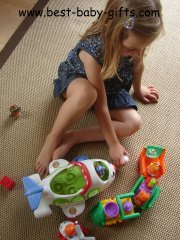 a little girl sitting on a carpet playing with a Little People plane and train